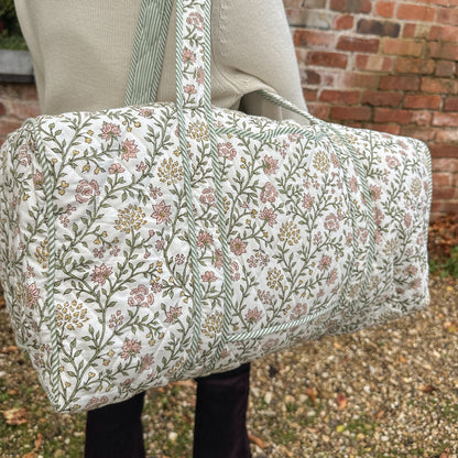 A person carrying a large quilted duffel bag with a floral print. The bag features a cream background with a delicate pattern of pink, green, and yellow flowers and stems, with striped green and white handles.