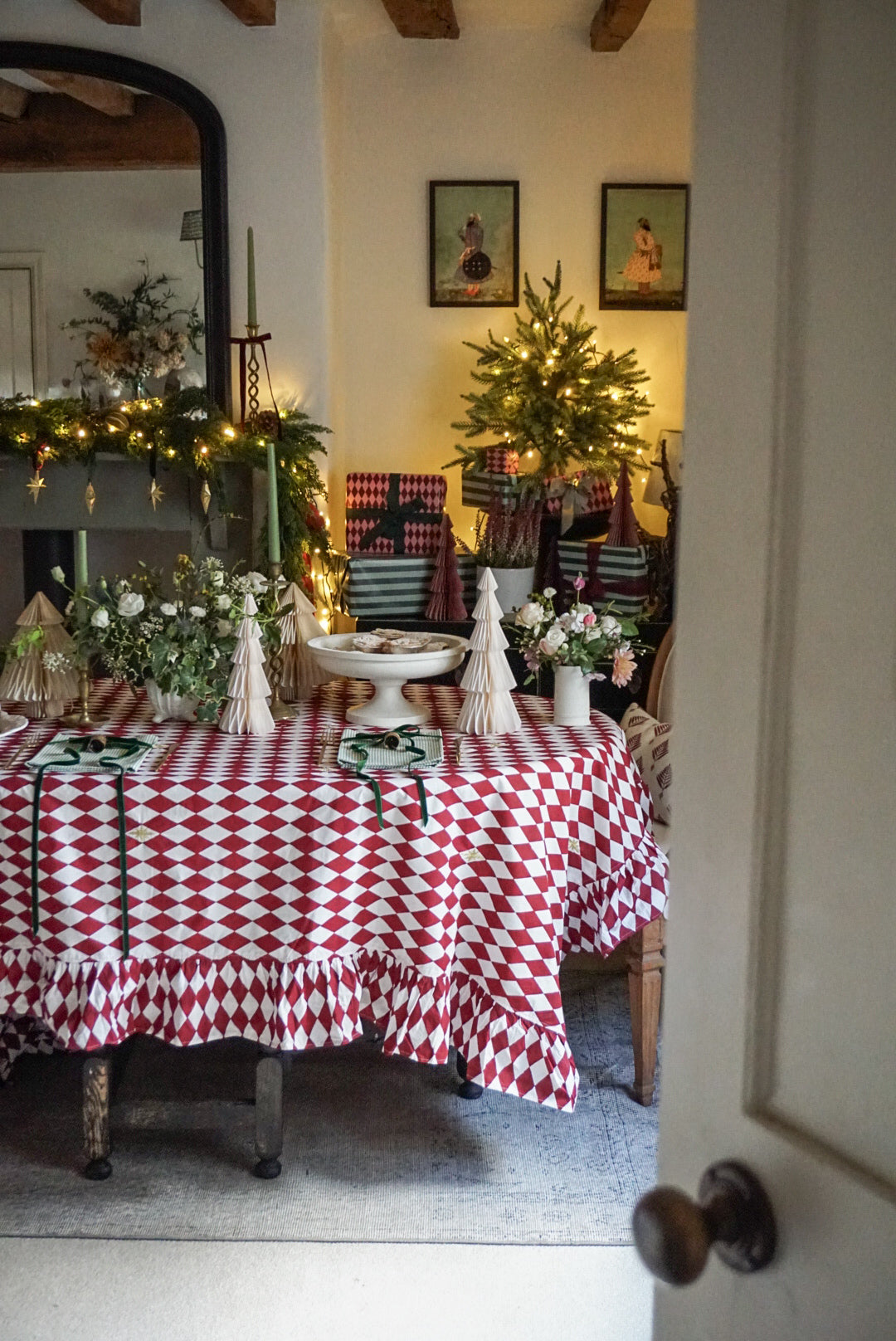 A festive holiday dining table decorated with a red and white diamond-patterned ruffled tablecloth, white cone trees, floral arrangements, and a white pedestal plate, set within a warmly lit home interior.